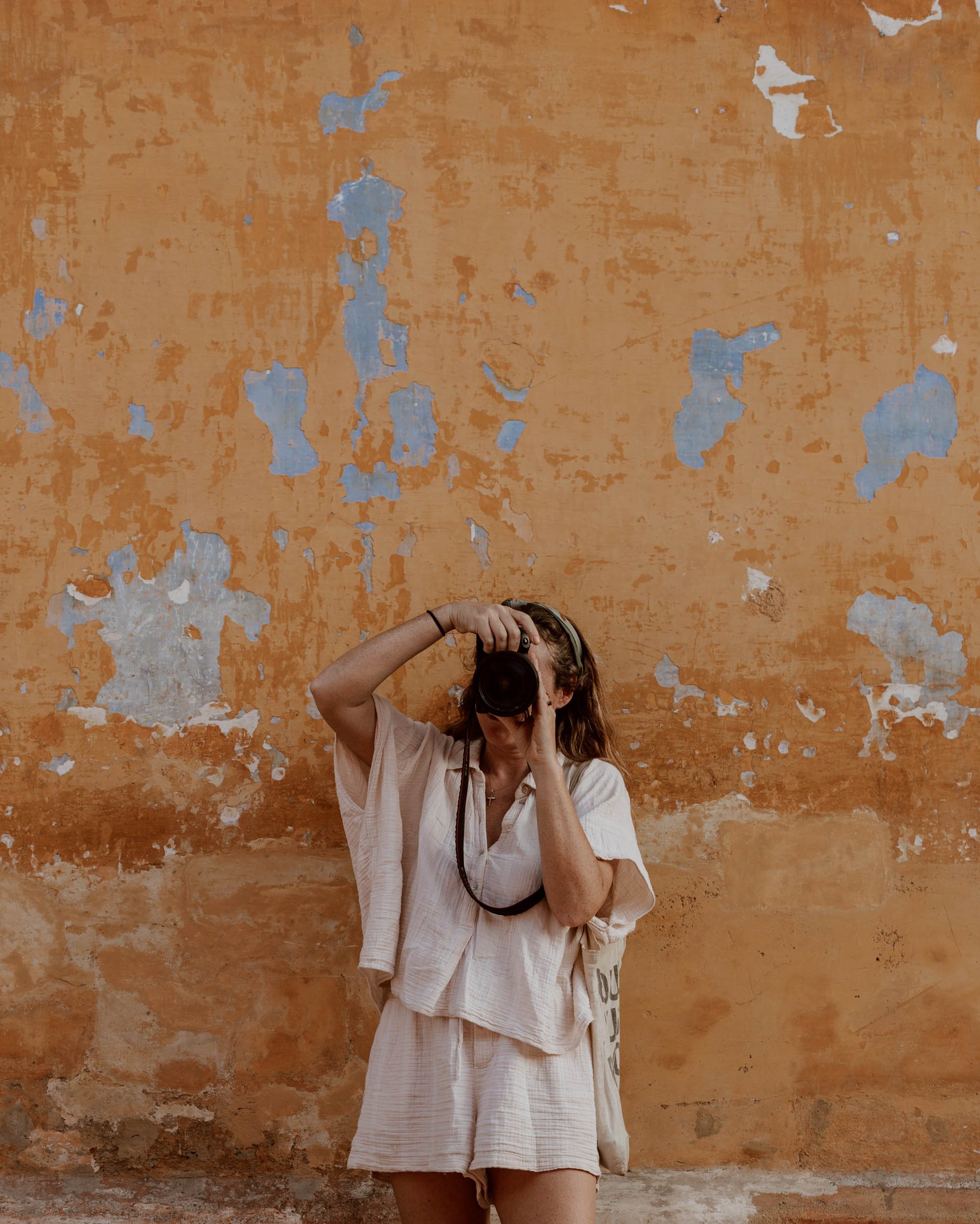 Person in light clothing photographing with camera against a weathered orange wall with blue paint peeling off.